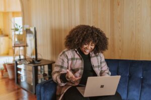 a woman of colour is working remotely on a laptop sitting on a blue sofa