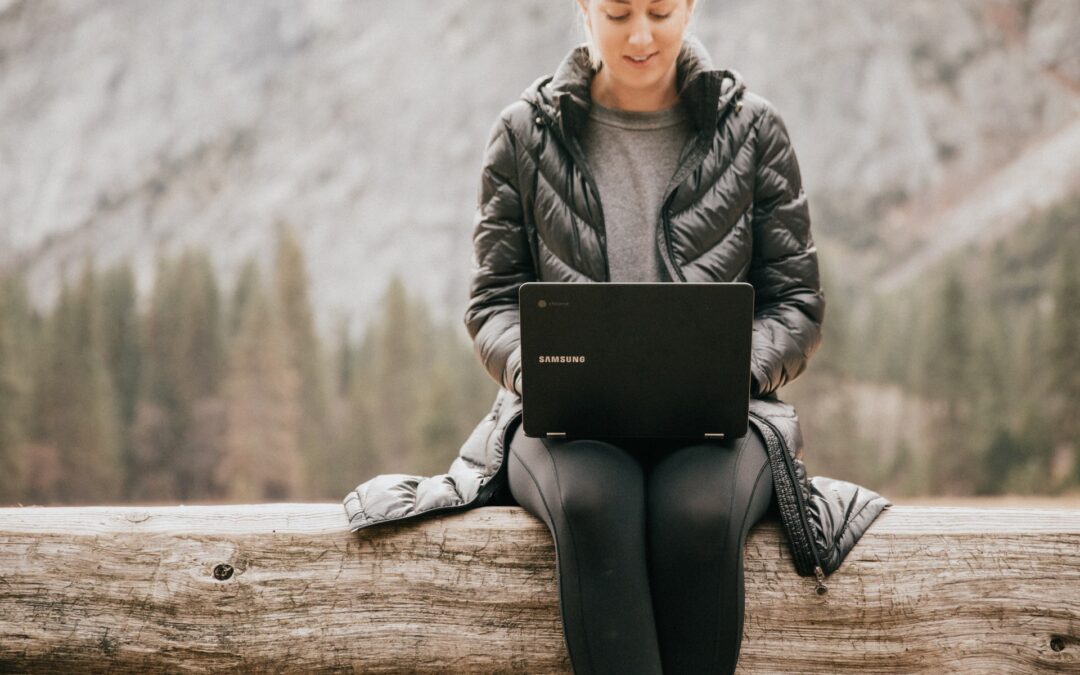 a woman working remotely outside sat on a tree trunk on a laptop